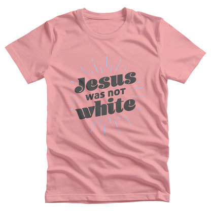 Pink unisex t-shirt that says, “Jesus was not white” in over a blue-green starburst. The text is thick, has curls, and is slanted upwards.