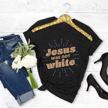 Black unisex t-shirt that says, “Jesus was not white” in over a blue-green starburst. The text is thick, has curls, and is slanted upwards.