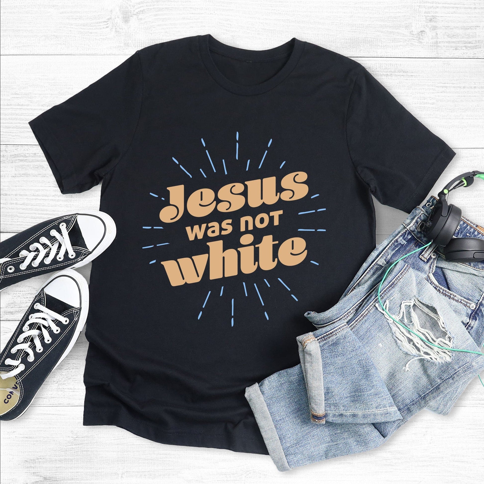 Black unisex t-shirt that says, “Jesus was not white” in over a blue-green starburst. The text is thick, has curls, and is slanted upwards.