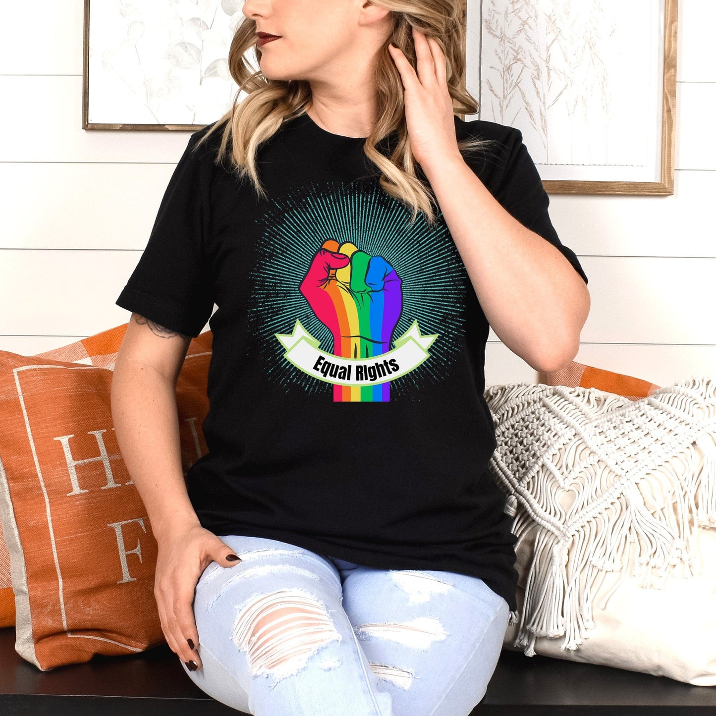 Black unisex t-shirt with a blue-green starburst and a rainbow fist in front of it. In a ribbon on the bottom of the fist over the wrist area, it says, “Equal Rights.”