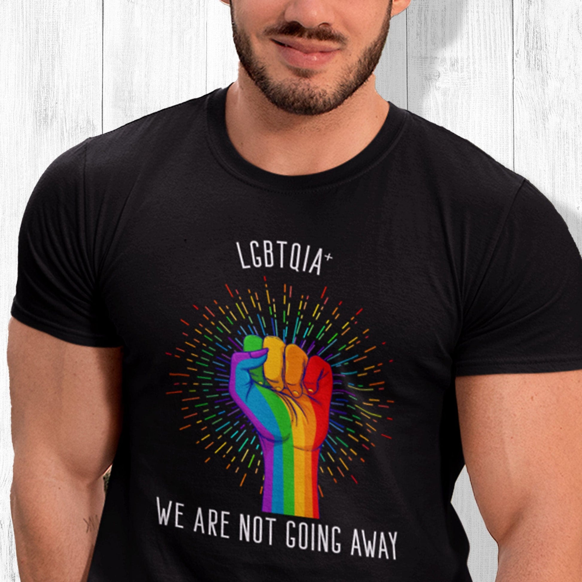 Black color unisex t-shirt that says, “LGBTQIA+ We Are Not Going Away” with a graphic of a rainbow fist with a rainbow-gradient color starburst behind it. Slightly arched over the starburst are the words “LGBTQIA+” and the rest of the text is below the graphic in one horizontal line.