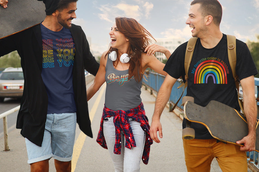 Three friends wearing LGBTQ+ shirts. The man on the left is wearing a t-shirt that says, "BE YOU" 5 times in rainbow colors in an outlined font. The woman in the middle is wearing a tank top that says, "BE YOU" in retro colors in a script font. The man on the right is waring a t-shirt that says, "My favorite color is rainbow" in all caps in rainbow colors, arched over a graphic of a rainbow.