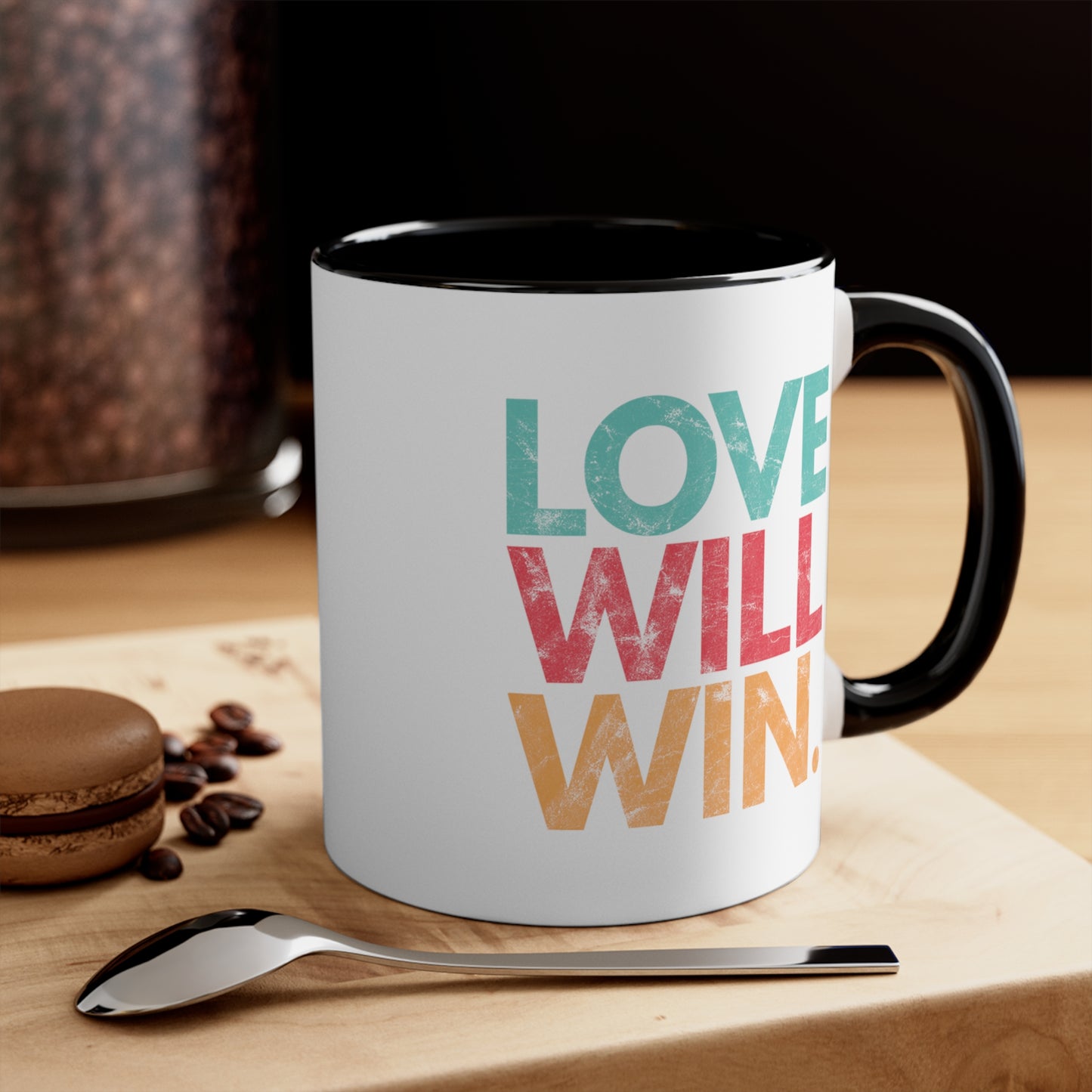 11oz ceramic mug with a black handle and interior with a graphic that says “Love will win.” in all caps. Each word is on its own line and is a different color. “Love” is greenish-blue, “Will” is red, and “Win.” is yellow. The graphic has a faded and slightly grungy/worn look. The mug is sitting on a wooden table with a spoon and a cookie.