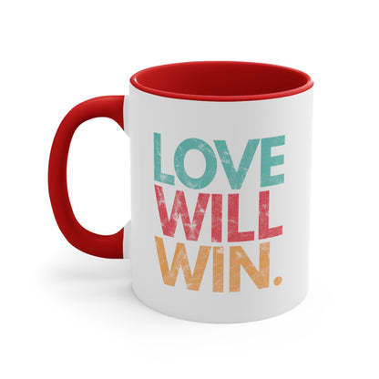 11oz ceramic mug with a red handle and interior with a graphic that says “Love will win.” in all caps. Each word is on its own line and is a different color. “Love” is greenish-blue, “Will” is red, and “Win.” is yellow. The graphic has a faded and slightly grungy/worn look.