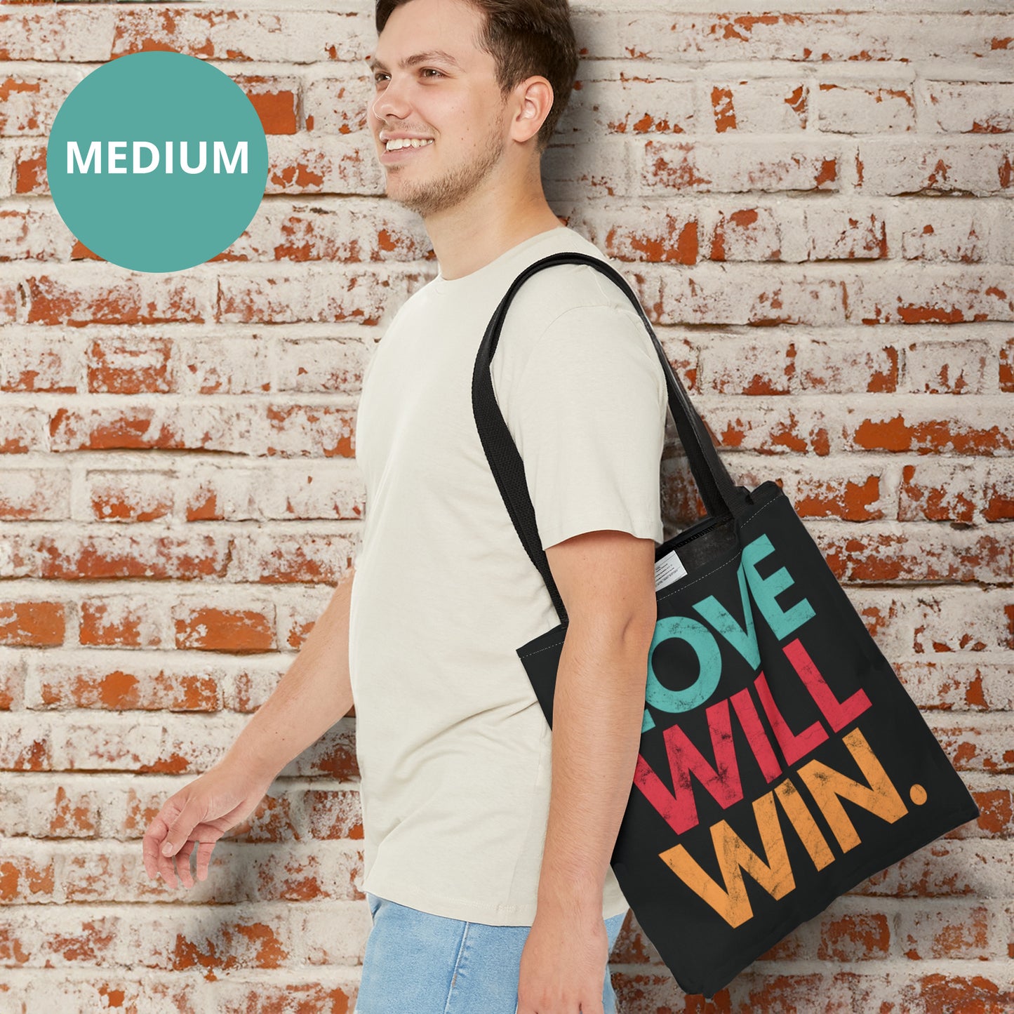 Black tote bag with a graphic that says “Love will win.” in all caps. Each word is on its own line and is a different color. “Love” is greenish-blue, “Will” is red, and “Win.” is yellow. The graphic has a faded and slightly grungy/worn look.