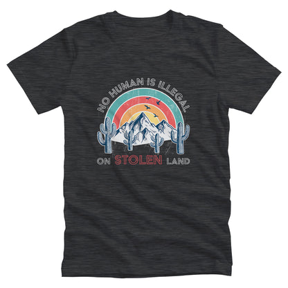 Dark Grey Heather color unisex t-shirt that says, “No Human is Illegal on Stolen Land” arched over a retro sunset with mountains and cactuses in the front. “No Human is Illegal” is arched over the top of the graphic and “on Stolen Land” is on one horizontal line beneath the graphic. The whole image is slightly distressed.