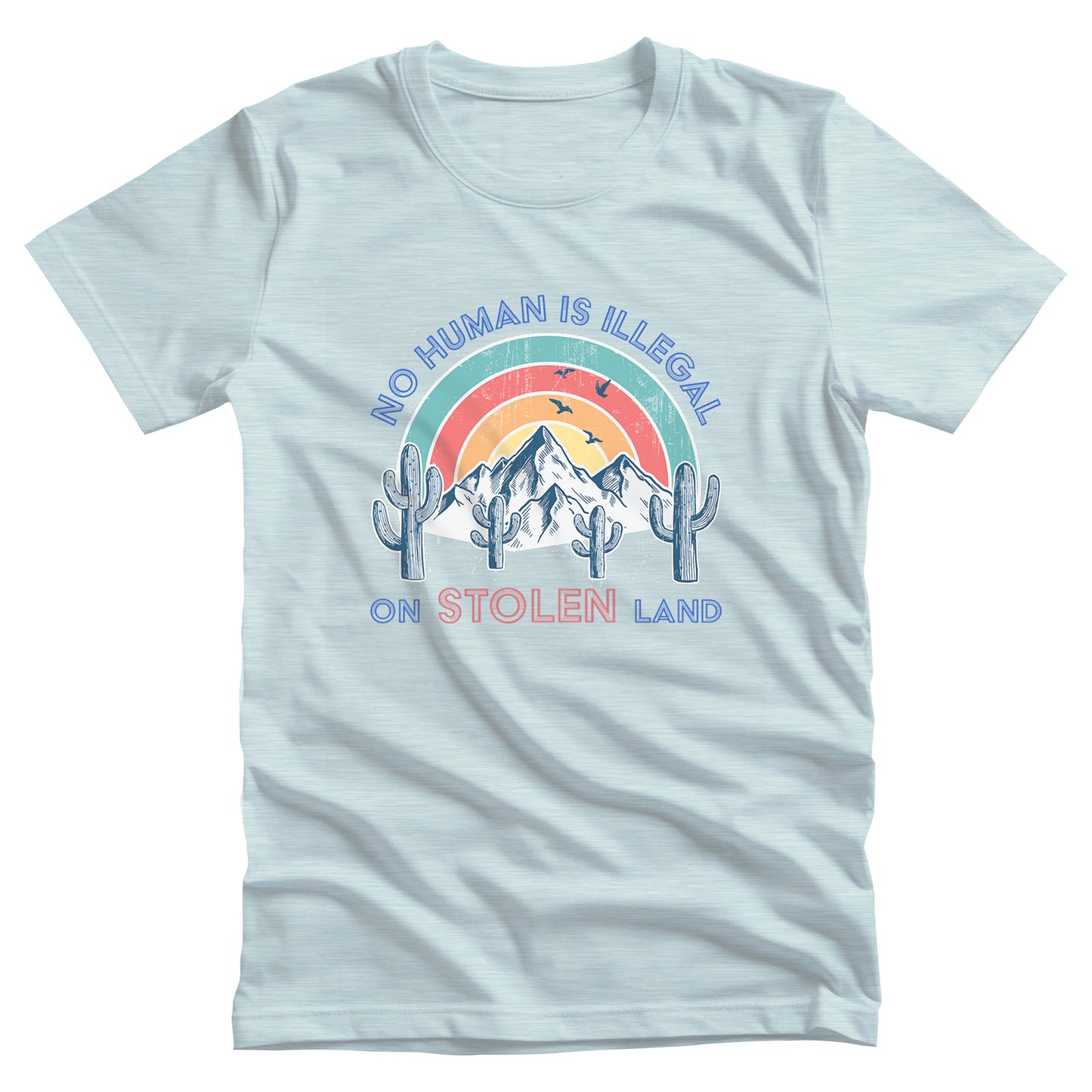 Heather Ice Blue color unisex t-shirt that says, “No Human is Illegal on Stolen Land” arched over a retro sunset with mountains and cactuses in the front. “No Human is Illegal” is arched over the top of the graphic and “on Stolen Land” is on one horizontal line beneath the graphic. The whole image is slightly distressed.