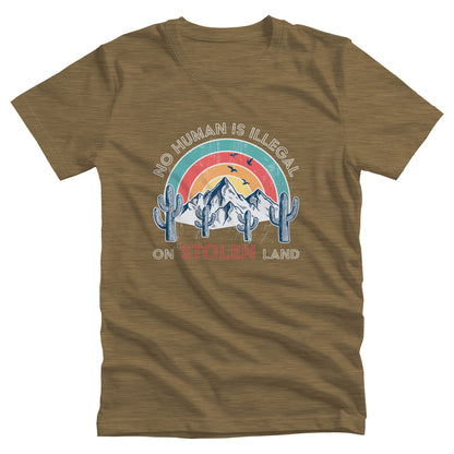 Heather Olive color unisex t-shirt that says, “No Human is Illegal on Stolen Land” arched over a retro sunset with mountains and cactuses in the front. “No Human is Illegal” is arched over the top of the graphic and “on Stolen Land” is on one horizontal line beneath the graphic. The whole image is slightly distressed.