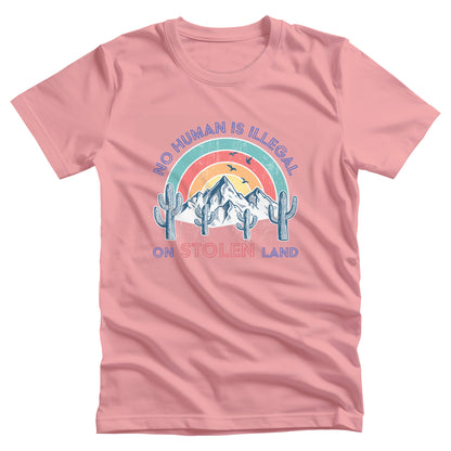 Pink unisex t-shirt that says, “No Human is Illegal on Stolen Land” arched over a retro sunset with mountains and cactuses in the front. “No Human is Illegal” is arched over the top of the graphic and “on Stolen Land” is on one horizontal line beneath the graphic. The whole image is slightly distressed.