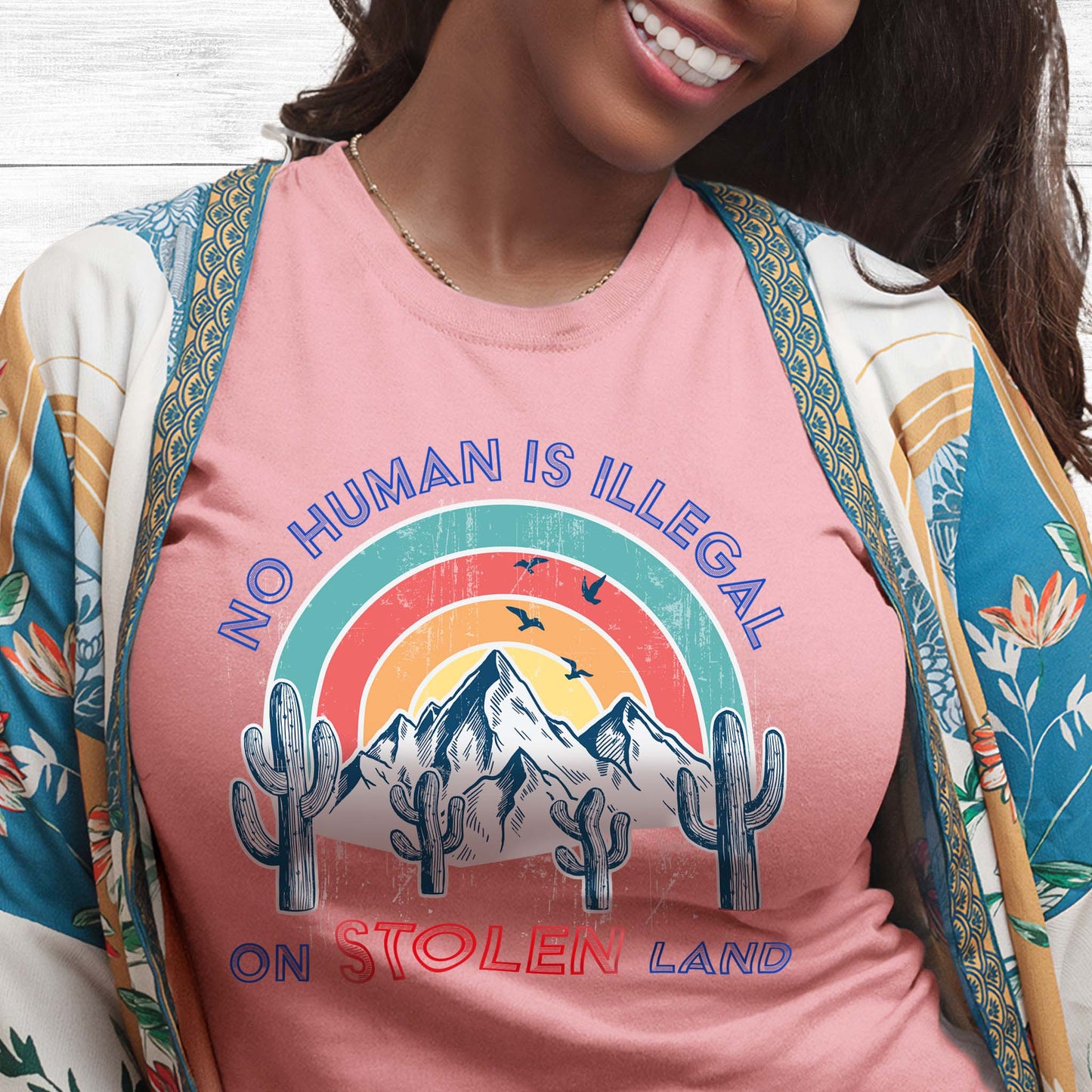 Pink unisex t-shirt that says, “No Human is Illegal on Stolen Land” arched over a retro sunset with mountains and cactuses in the front. “No Human is Illegal” is arched over the top of the graphic and “on Stolen Land” is on one horizontal line beneath the graphic. The whole image is slightly distressed.