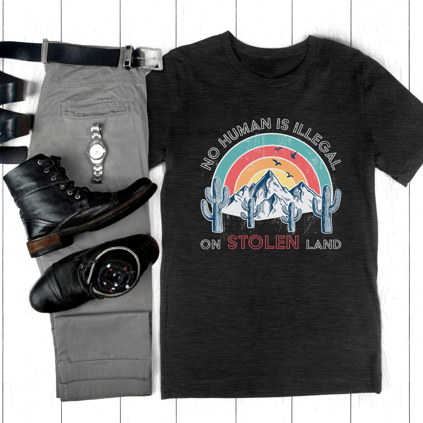 Black unisex t-shirt that says, “No Human is Illegal on Stolen Land” arched over a retro sunset with mountains and cactuses in the front. “No Human is Illegal” is arched over the top of the graphic and “on Stolen Land” is on one horizontal line beneath the graphic. The whole image is slightly distressed.