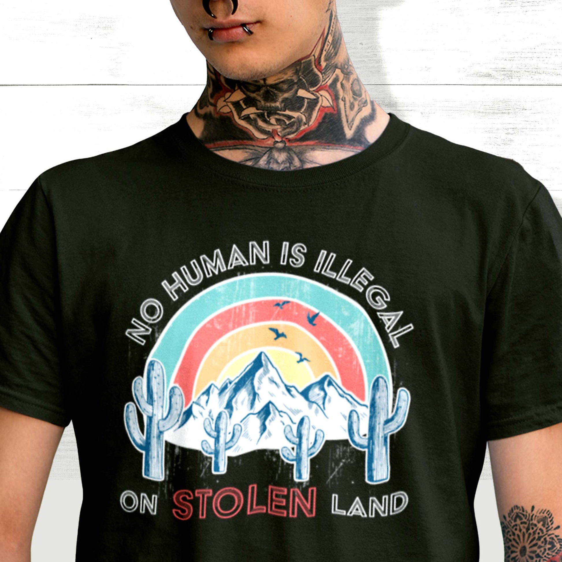 Black unisex t-shirt that says, “No Human is Illegal on Stolen Land” arched over a retro sunset with mountains and cactuses in the front. “No Human is Illegal” is arched over the top of the graphic and “on Stolen Land” is on one horizontal line beneath the graphic. The whole image is slightly distressed.