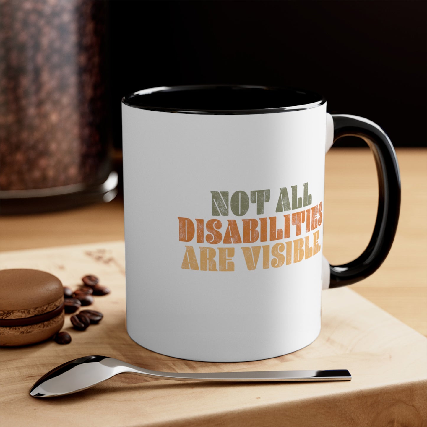 Contextual photo of an 11oz ceramic mug with a black interior and handle that says, “Not all disabilities are visible” in a thick, retro font in all caps. Each line is a different color. “Not all” is light green, “disabilities” is orange, and “are visible” is yellow. The graphic has a retro look and is slightly distressed. The mug is on a table with a spoon and a cookie.