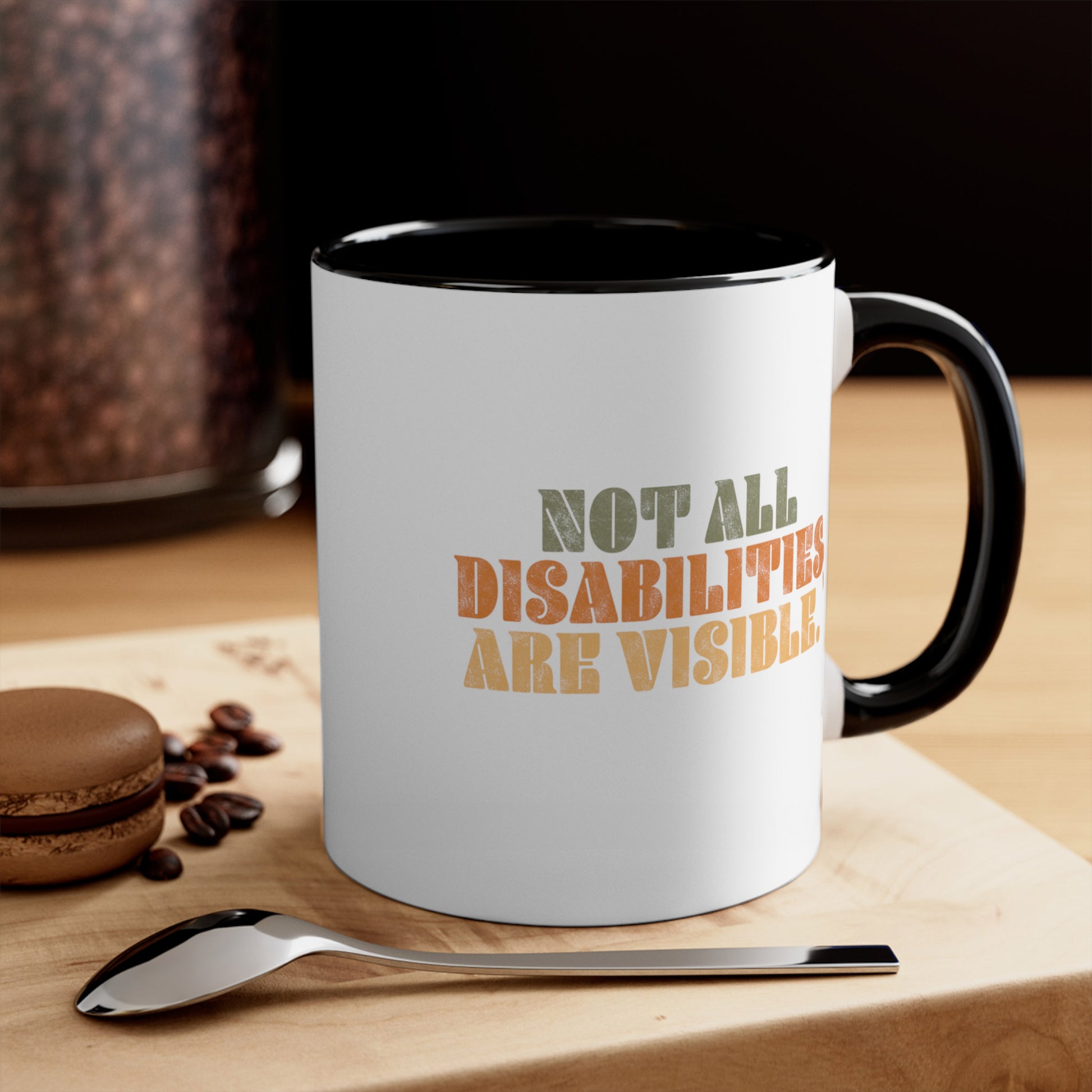 Contextual photo of an 11oz ceramic mug with a black interior and handle that says, “Not all disabilities are visible” in a thick, retro font in all caps. Each line is a different color. “Not all” is light green, “disabilities” is orange, and “are visible” is yellow. The graphic has a retro look and is slightly distressed. The mug is on a table with a spoon and a cookie.