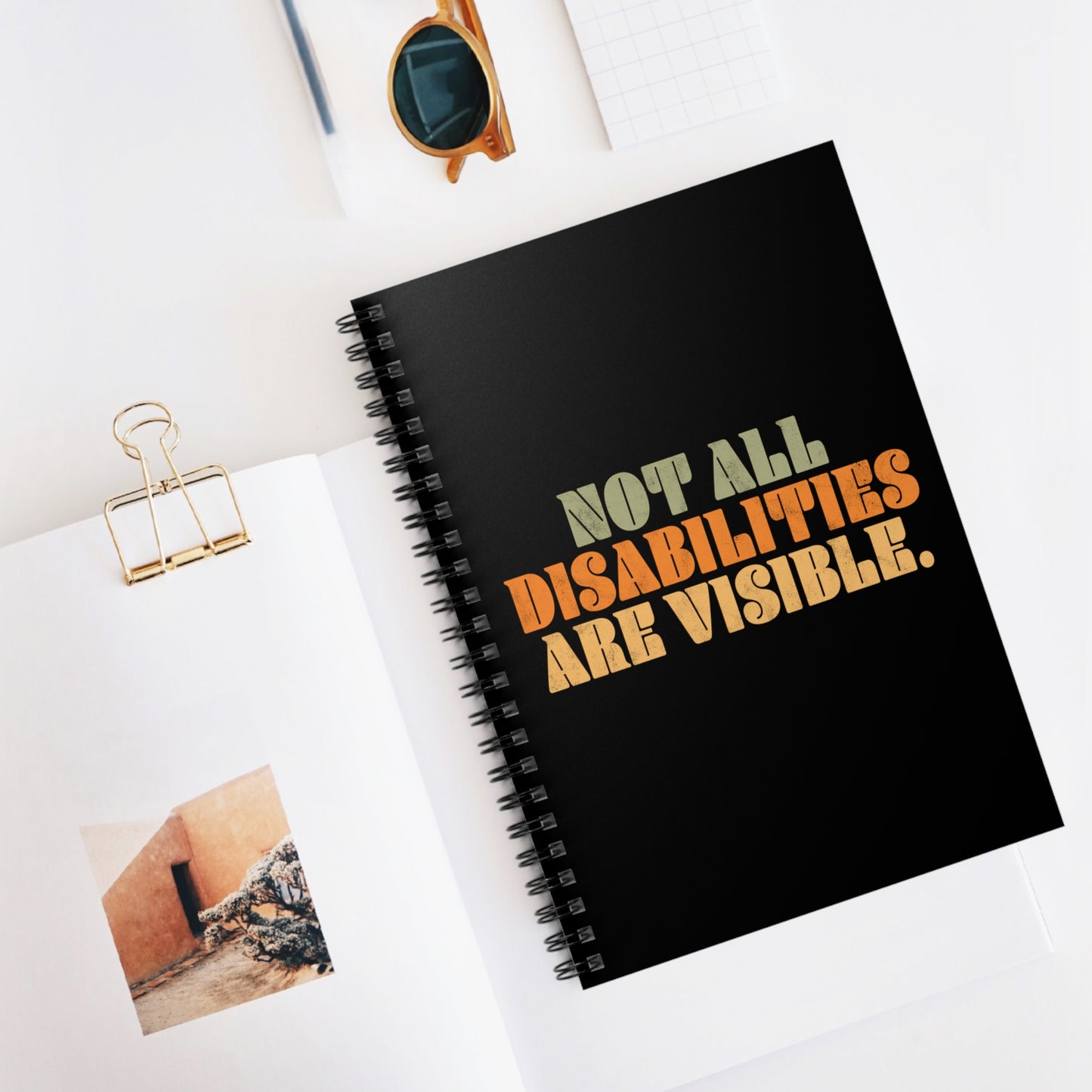 Contextual photo of a black spiral notebook that says, “Not all disabilities are visible” in a thick, retro font in all caps. Each line is a different color. “Not all” is light green, “disabilities” is orange, and “are visible” is yellow. The graphic has a retro look and is slightly distressed. The mug is on a table with a spoon and a cookie.