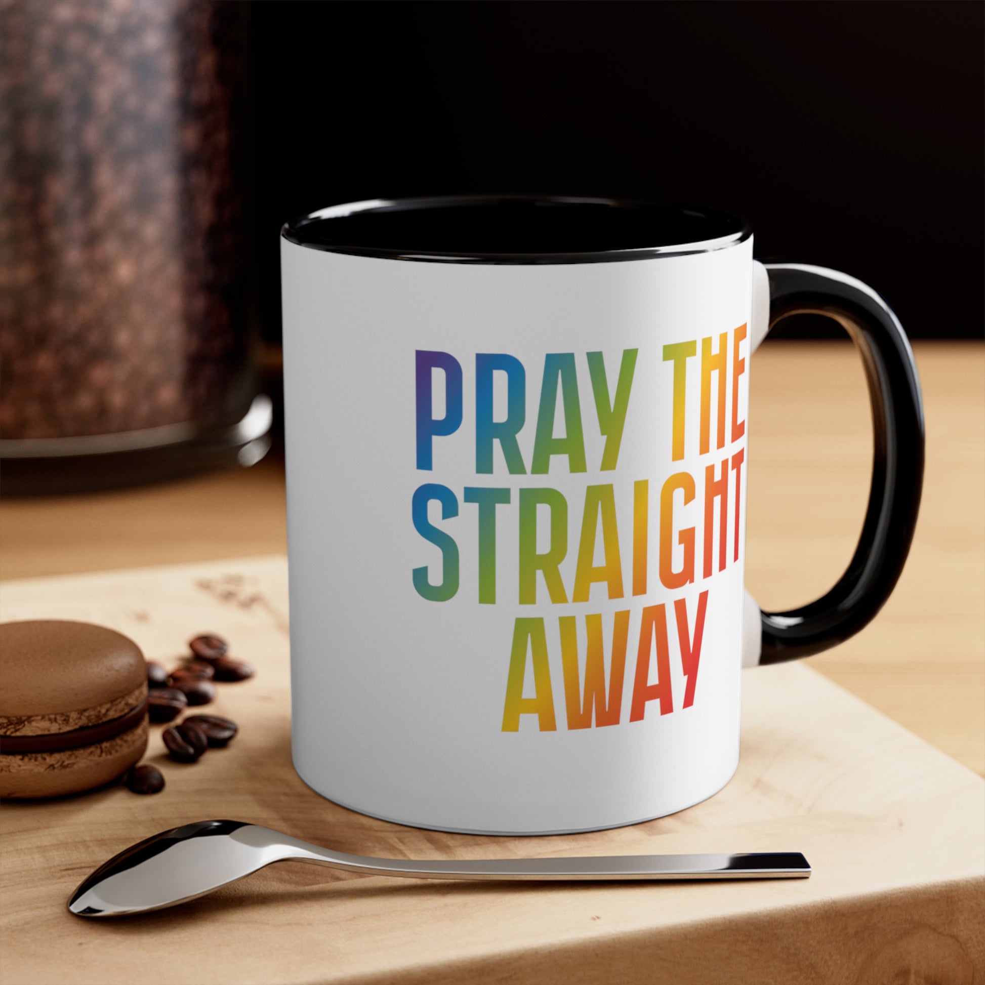 Contextual photo of an 11oz ceramic mug with a black interior and handle that says, “PRAY THE STRAIGHT AWAY” in a bold font with gradient rainbow colors. 