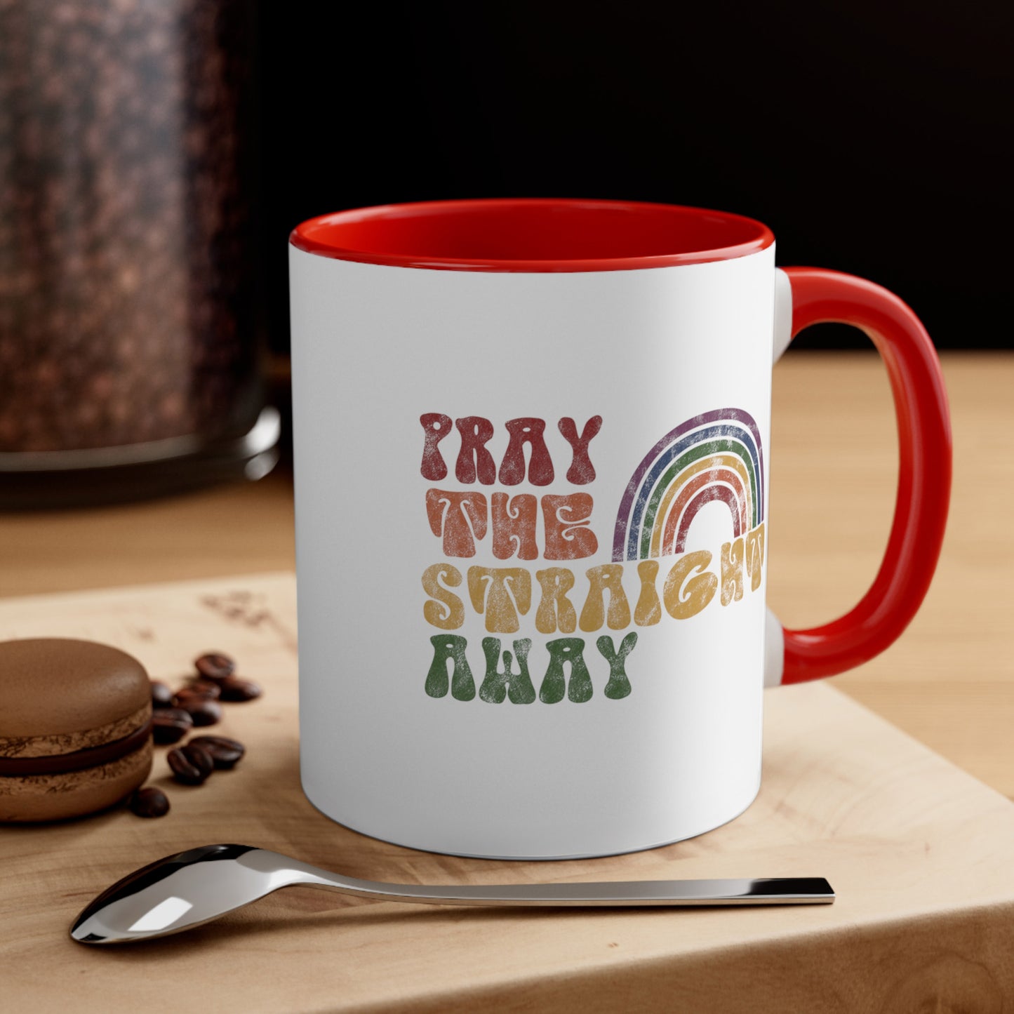 Contextual photo of an 11oz ceramic mug with a red interior and handle that says, “PRAY THE STRAIGHT AWAY” in a retro font. Each word is a different retro color and on its own line. “PRAY” is maroon, “THE” is orange, “STRAIGHT” is yellow, and “AWAY” is green. The words are stacked and left-aligned with a rainbow graphic that has matching colors nestled after the word “THE” over the word “STRAIGHT.”