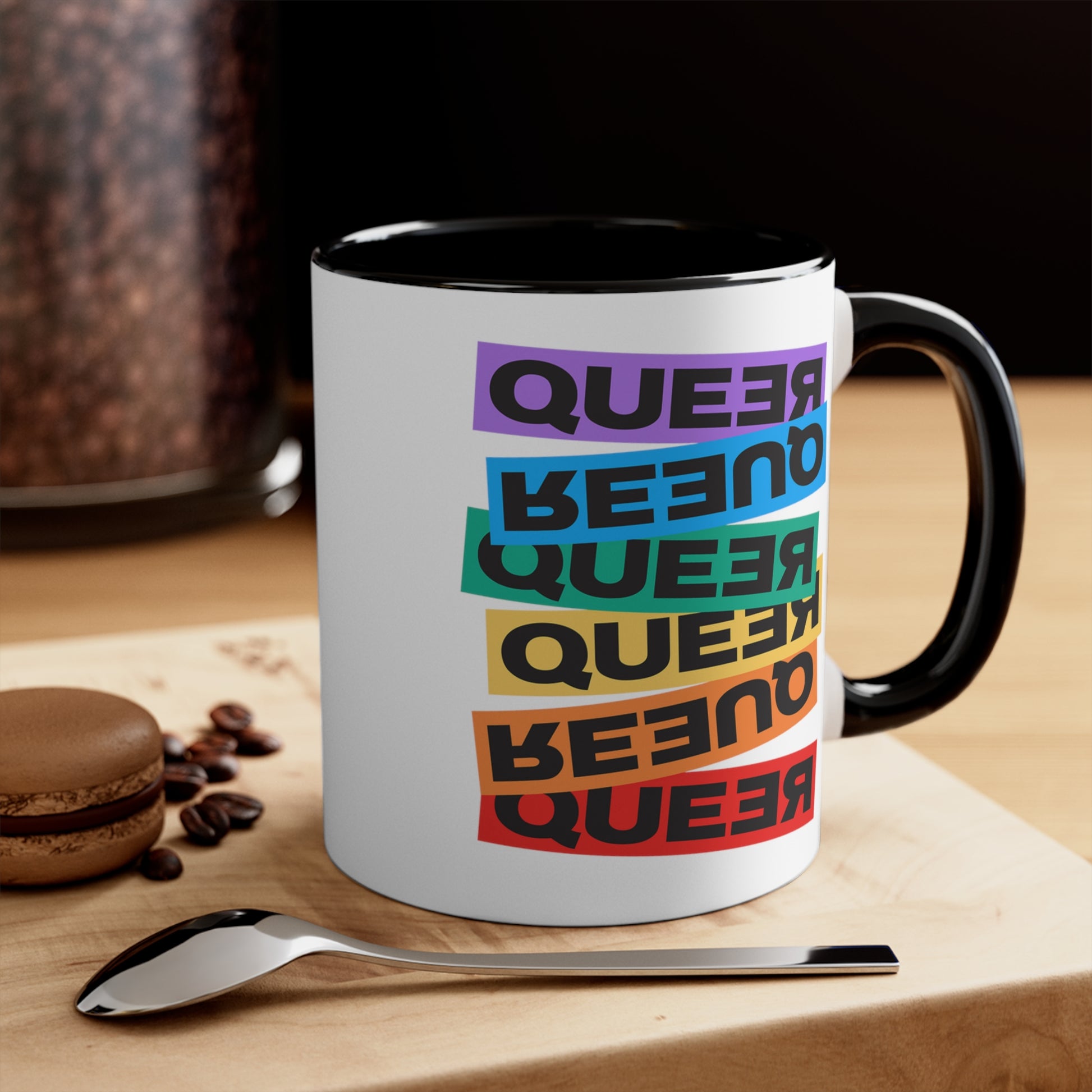 Contextual image of an 11oz ceramic mug with a black handle and interior. This mug artistically expresses the word “Queer.” Each variation of the word is black text in a rectangular block and stacked vertically at a slightly different angle than the next row/block. Sometimes “Queer” is backward, sometimes upside down, and sometimes 1-2 letters are backward. The color blocks are purple, blue, green, yellow, and red.