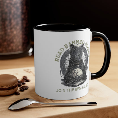 11oz ceramic mug with a black interior and handle with a graphic of a cat sitting over a globe and an open book. The text says “Read banned books” arched over the top of the cat and “Join the resistance” in a line under the graphic. The text is in all caps. This image is contextual with the mug sitting on a wooden table with a spoon and a cookie beside it.