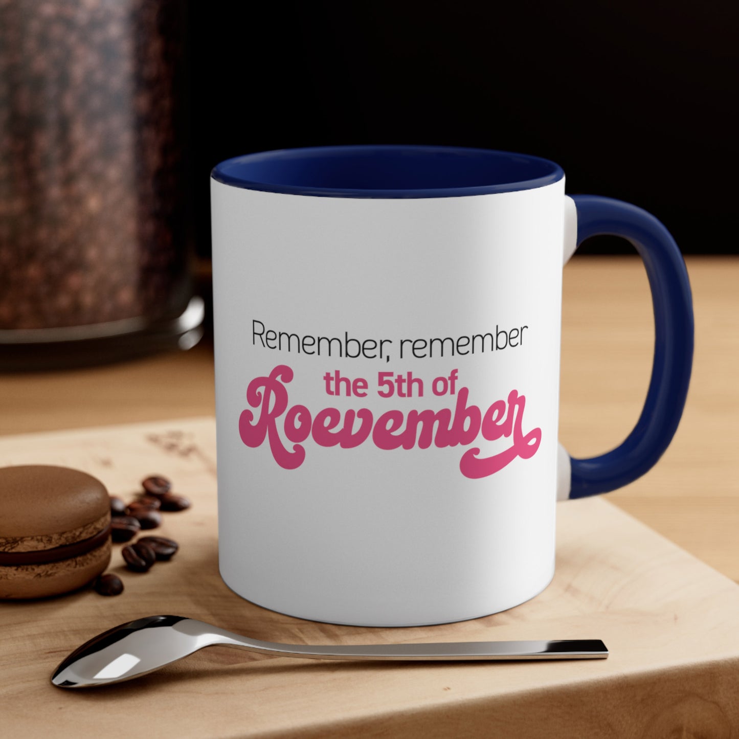 Contextual image of an 11oz ceramic mug with a navy interior and handle featuring the phrase 'Remember, Remember the 5th of Roevember'. The text 'The 5th of Roevember' is highlighted in pink, embellished with a decorative script font and additional swooshes. The mug is on a table with a spoon and a cookie.