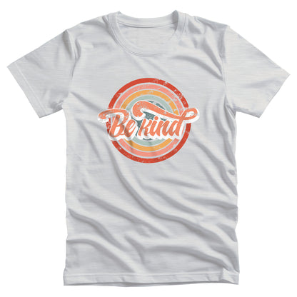 Ash color unisex t-shirt with a graphic that says “Be Kind.” There is a retro-colored circular rainbow behind it. The text is outlined in white and has a long swoosh that swirls from the end of the letter “d” across the entire word “kind.”