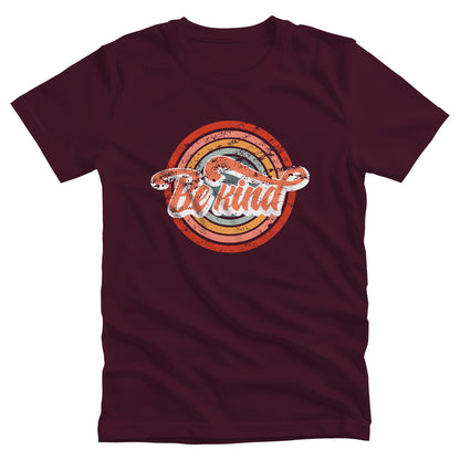 Maroon unisex t-shirt with a graphic that says “Be Kind.” There is a retro-colored circular rainbow behind it. The text is outlined in white and has a long swoosh that swirls from the end of the letter “d” across the entire word “kind.”