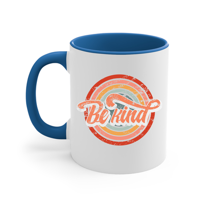 11oz white ceramic mug with a blue handle and interior that says “Be Kind.” There is a retro-colored circular rainbow behind it. The text is outlined in white and has a long swoosh that swirls from the end of the letter “d” across the entire word “kind.”