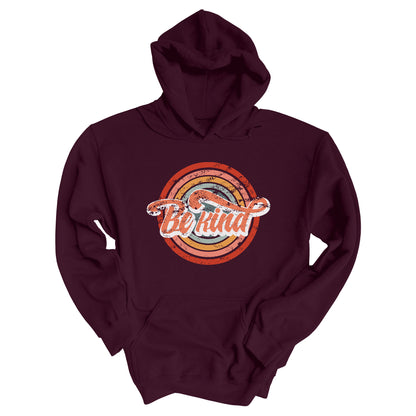 Maroon unisex hoodie with a graphic that says “Be Kind.” There is a retro-colored circular rainbow behind it. The text is outlined in white and has a long swoosh that swirls from the end of the letter “d” across the entire word “kind.”