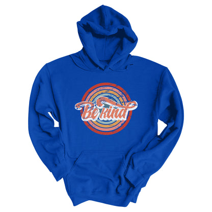 Royal blue unisex hoodie with a graphic that says “Be Kind.” There is a retro-colored circular rainbow behind it. The text is outlined in white and has a long swoosh that swirls from the end of the letter “d” across the entire word “kind.”