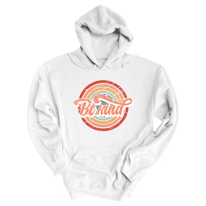 White unisex hoodie with a graphic that says “Be Kind.” There is a retro-colored circular rainbow behind it. The text is outlined in white and has a long swoosh that swirls from the end of the letter “d” across the entire word “kind.”