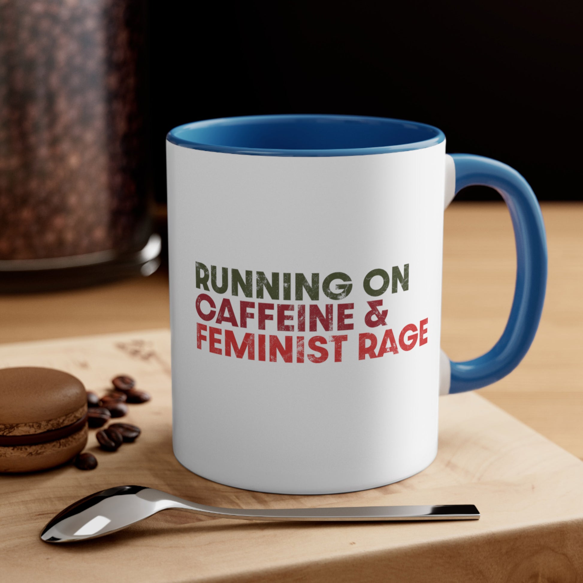 Contextual image of an 11oz ceramic mug with a blue interior and handle color that says, “RUNNING ON CAFFEINE & FEMINIST RAGE” with each line being a different color. “RUNNING ON” is dark green, “CAFFEINE &” is dark red, and “FEMINIST RAGE” is dark orange. The text is slightly distressed.