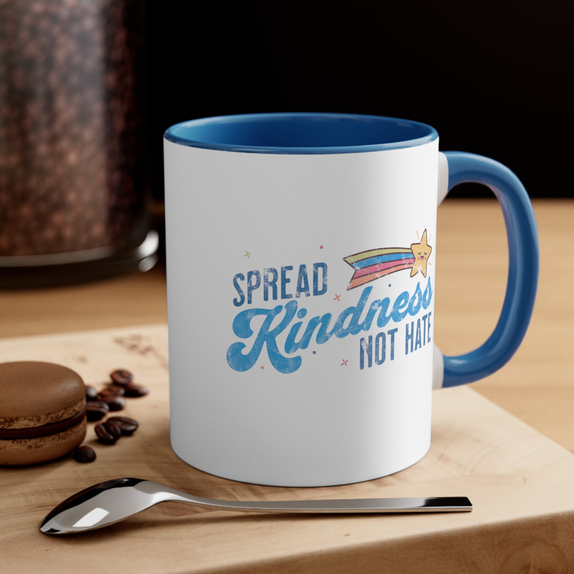 11oz ceramic mug with a blue interior and handle. It has a graphic that says “Spread Kindness Not Hate” with an illustration of a rainbow shooting star over the word “kindness.” That word is in a script font, and the rest is a regular font in all caps. ”Spread” is on top of the word “Kindness” and aligned left, while “Not Hate” is below the word “kindness” and aligned to the right. The mug is sitting on a table with a spoon and cookie in the picture.