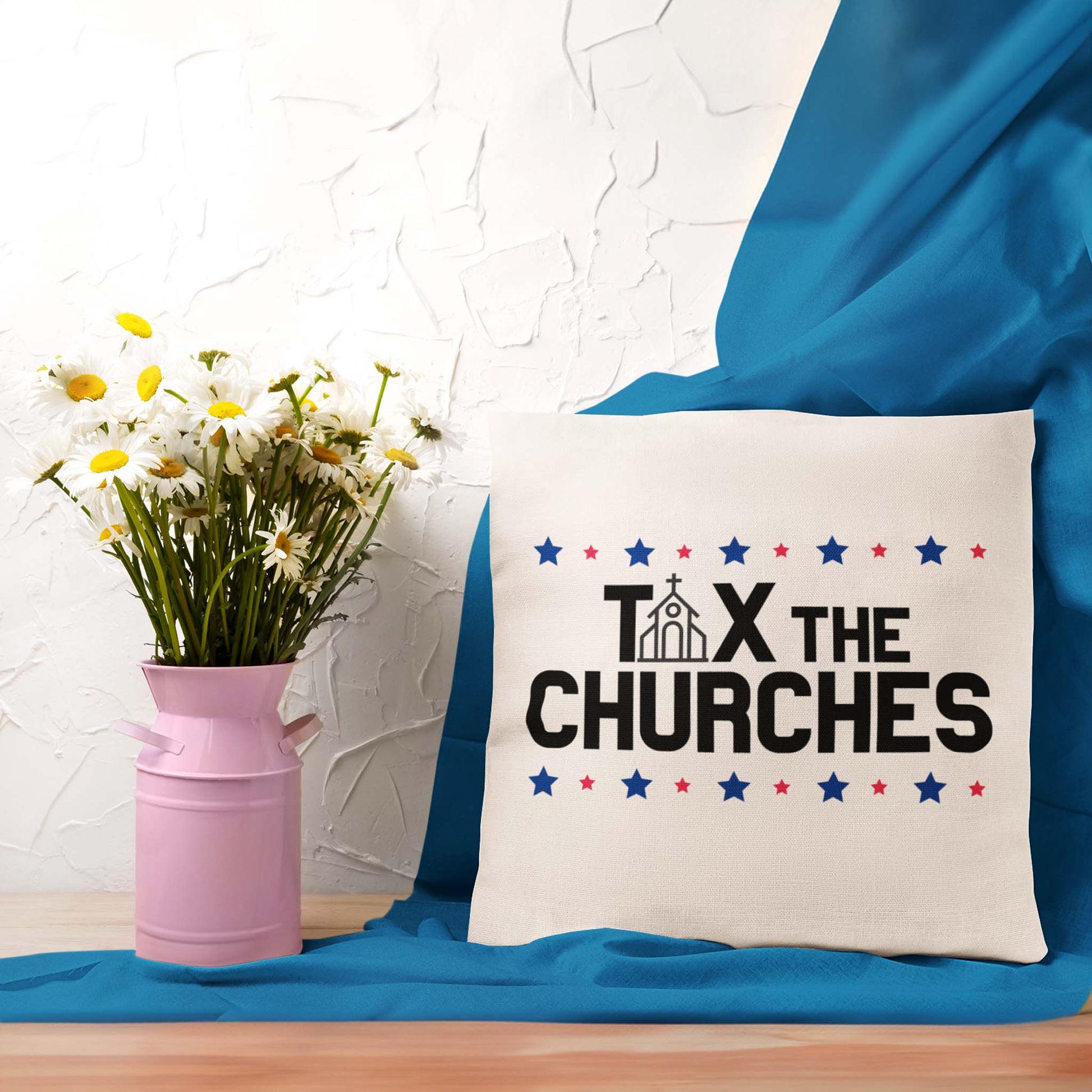 Contextual image of a beige color indoor square pillow featuring bold text that reads 'TAX THE CHURCHES,' with the letter 'A' in 'TAX' depicted as a church illustration. Alternating blue and red stars decorate the phrase above and below. There is a flower base and a blue blanket behind the pillow.