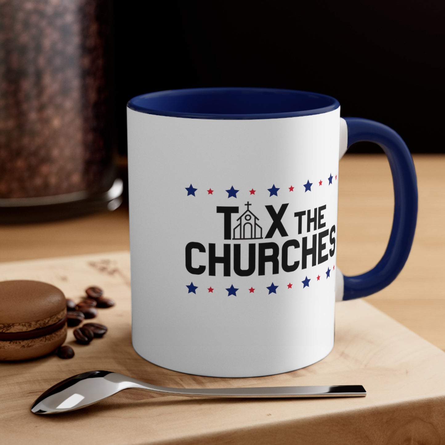 Contextual image of an 11oz ceramic mug with a navy interior and handle featuring bold text that reads 'TAX THE CHURCHES,' with the letter 'A' in 'TAX' depicted as a church illustration. Alternating blue and red stars decorate the phrase above and below. The mug is on a table with a spoon and a cookie.