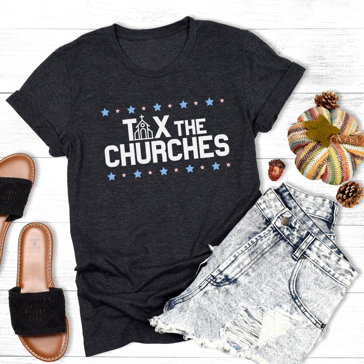 Dark Grey Heather color unisex t-shirt that says, “Tax the Churches” in all caps. There are alternating large and small stars in a line above and below the text. The “A” in “Tax” is a line illustration of a church.