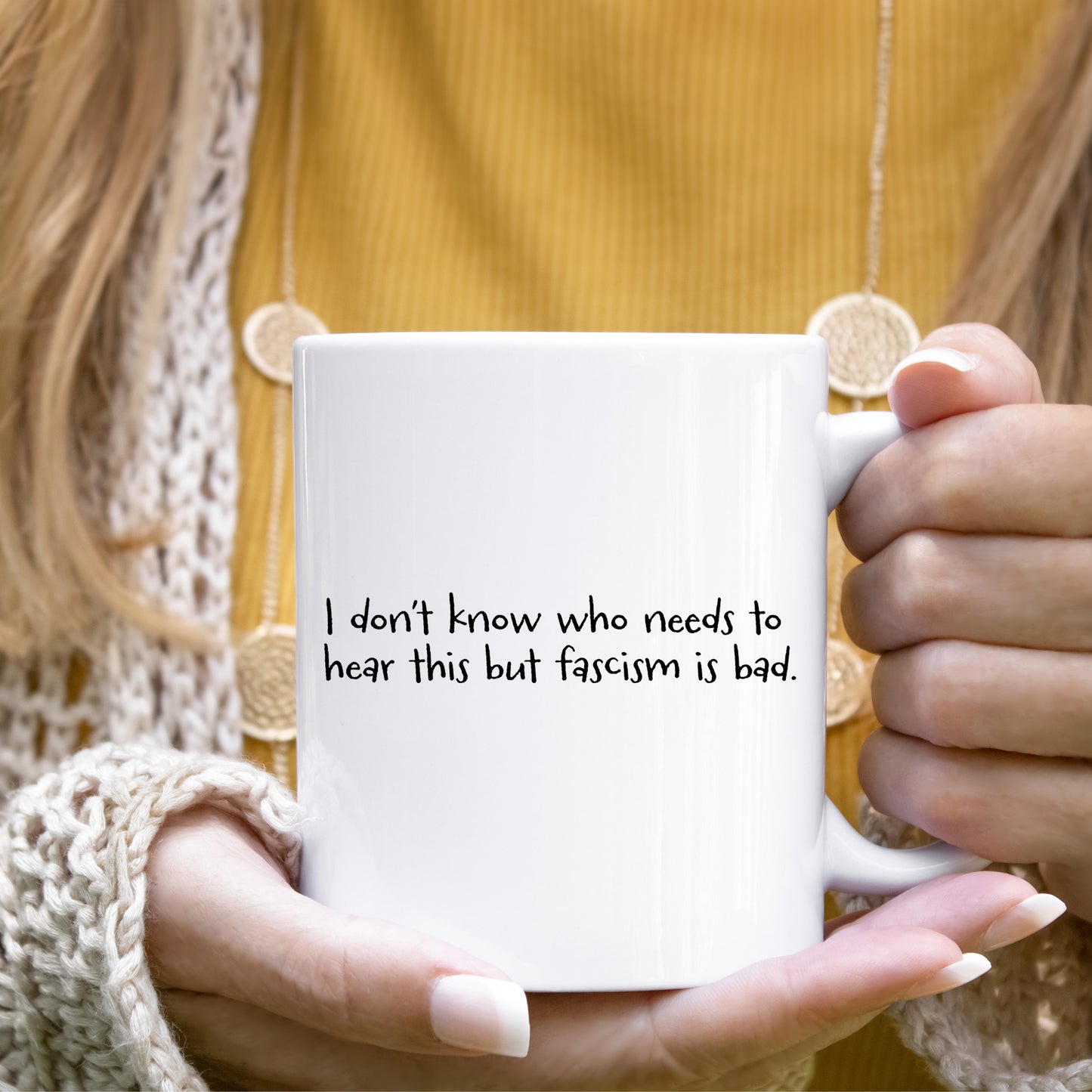 A woman holding a white ceramic mug with the handle facing the right that reads "I don't know who needs to hear this but fascism is bad." The text is written in a handwritten font.