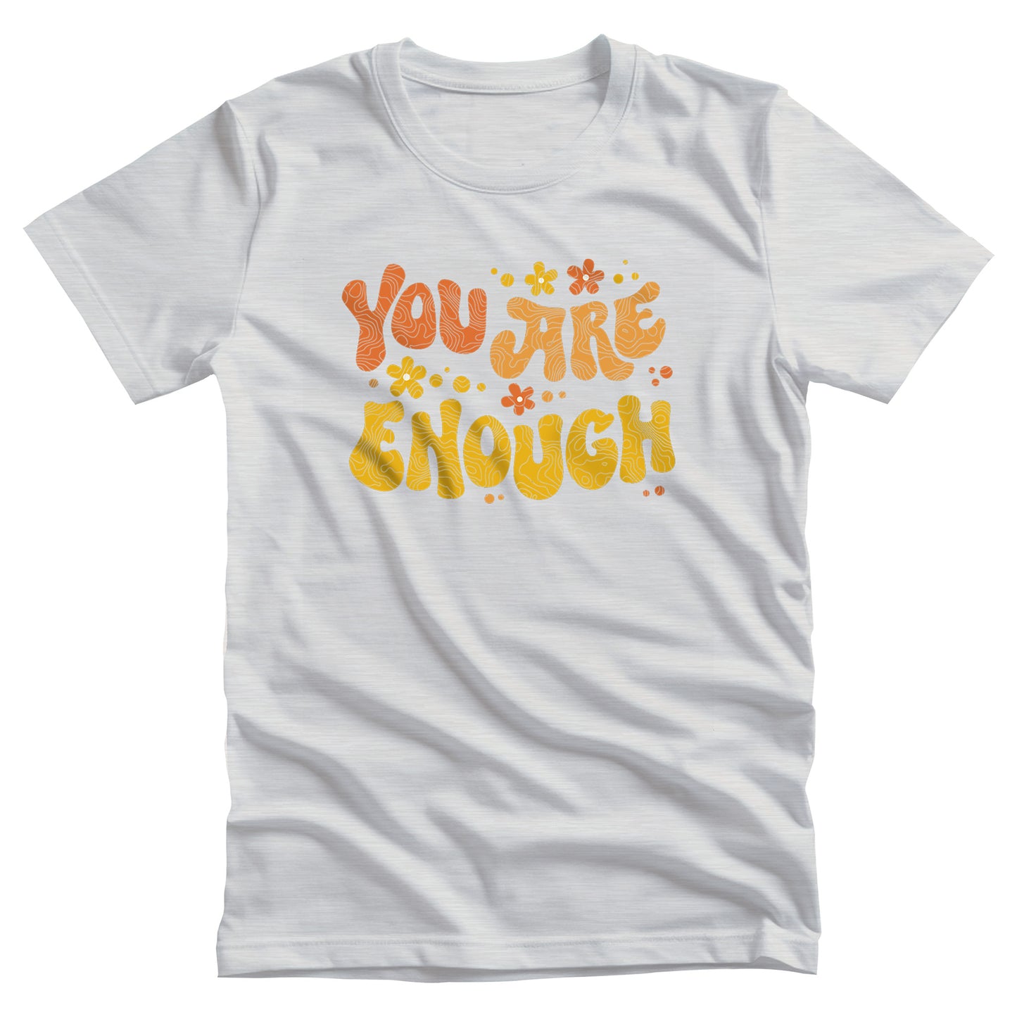 Ash color unisex t-shirt with a graphic that says “You Are Enough” in a retro font. There are small retro flowers spaced throughout. “You” is reddish-orange, “Are” is orange, and “Enough” is yellow, all retro as well.
