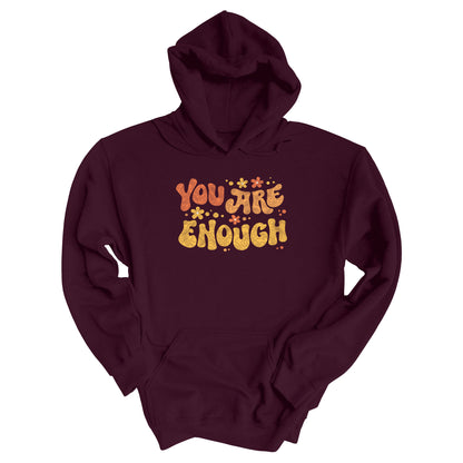 Maroon unisex liberal and progressive hoodie with a graphic that says “You Are Enough” in a retro font. There are small retro flowers spaced throughout. “You” is reddish-orange, “Are” is orange, and “Enough” is yellow, all retro as well.