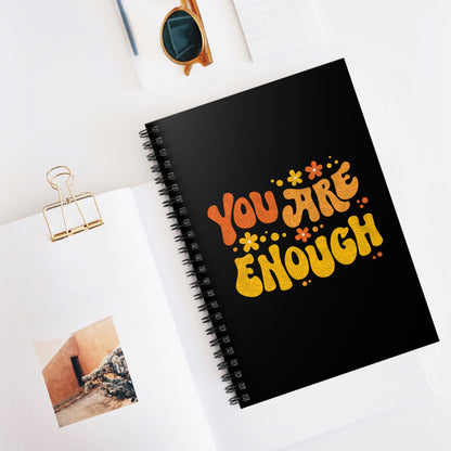 Contextual photo of a black spiral-bound notebook with a graphic on the front that says “You Are Enough” in a retro font. There are small retro flowers spaced throughout. “You” is reddish-orange, “Are” is orange, and “Enough” is yellow, all retro as well.