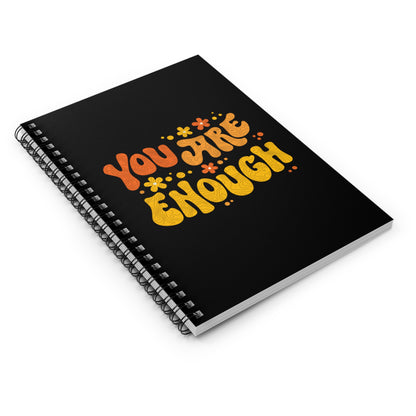 A black spiral-bound notebook with a graphic on the front that says “You Are Enough” in a retro font. There are small retro flowers spaced throughout. “You” is reddish-orange, “Are” is orange, and “Enough” is yellow, all retro as well.